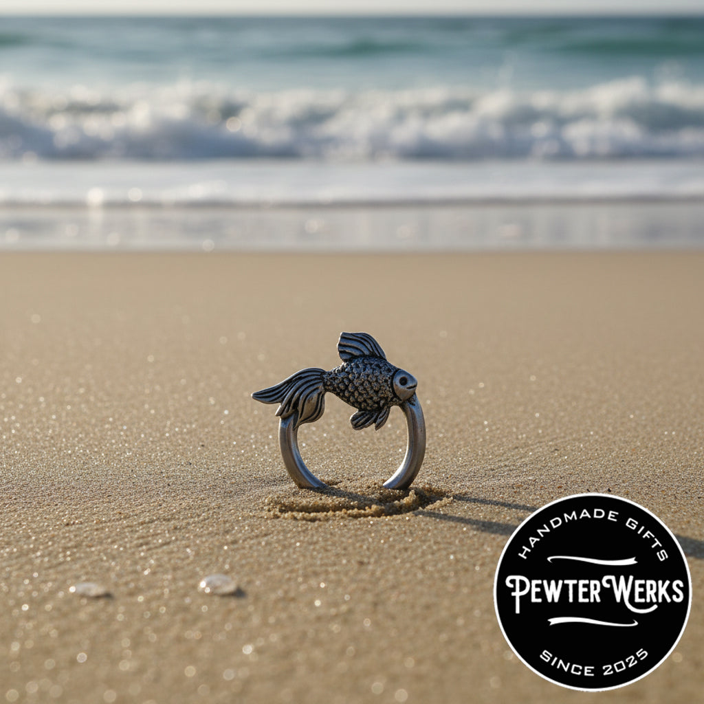 A Pewter 'Angelfish' Eyeglass Holder washed up on the beach in the sand. PewterWerks logo visible.