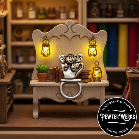 Pewter 'Angel in a Teacup' Eyeglass Holder sitting on a bench with some books and lanterns. PewterWerks logo visible.