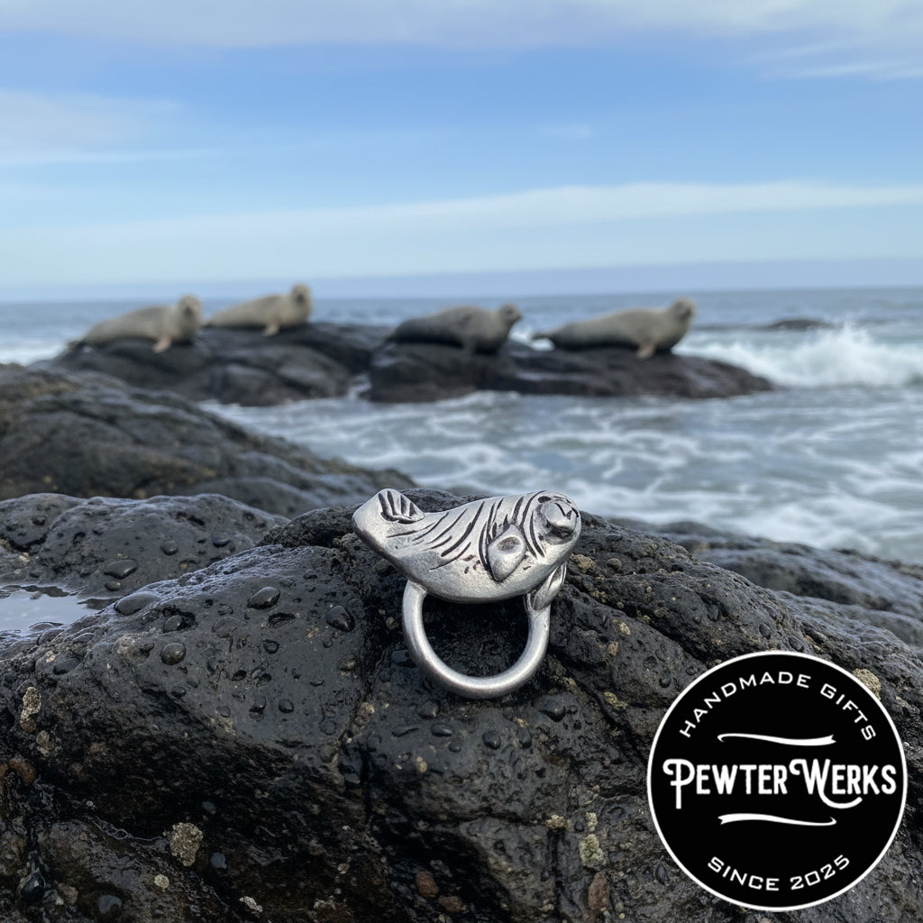 A pewter 'Baby Seal' eyeglass holder laying on a rock near the ocean with other seals in the background. PewterWerks logo visible.