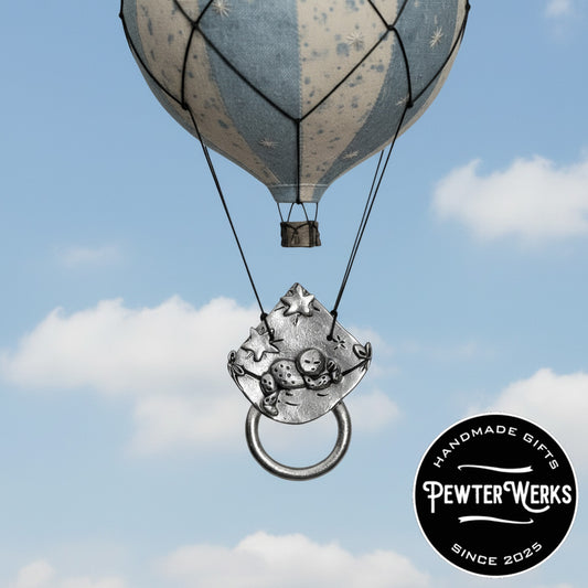 A Pewter 'Baby on a Blanket' Eyeglass Holder floating in the sky from a hot air balloon. PewterWerks logo visible.