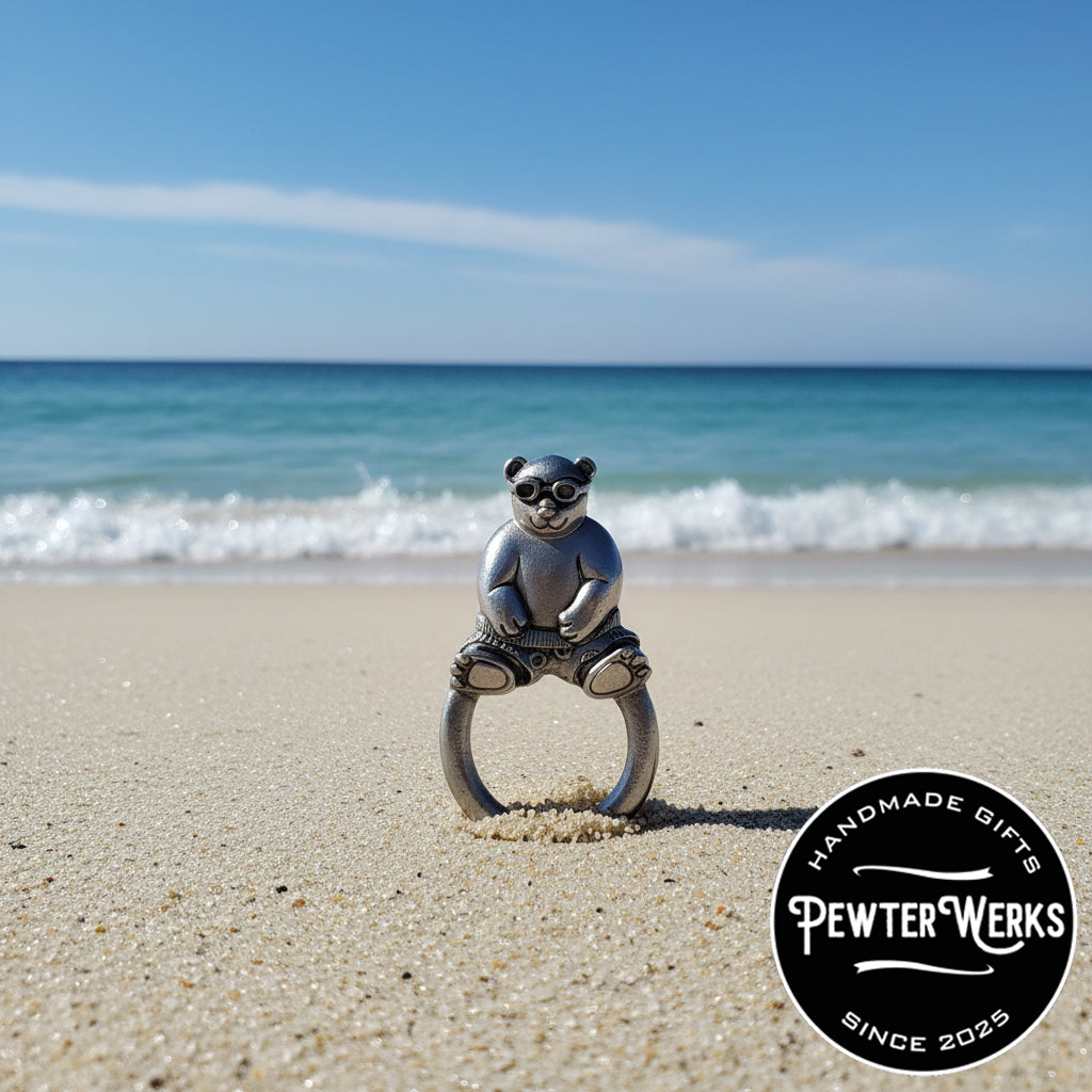 A pewter 'Beach Panda' eyeglass holder washed up on the beach in the sand with the ocean in the background. PewterWerks logo visible.