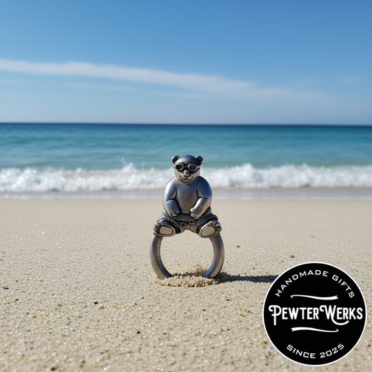 A pewter 'Beach Panda' eyeglass holder washed up on the beach in the sand with the ocean in the background. PewterWerks logo visible.