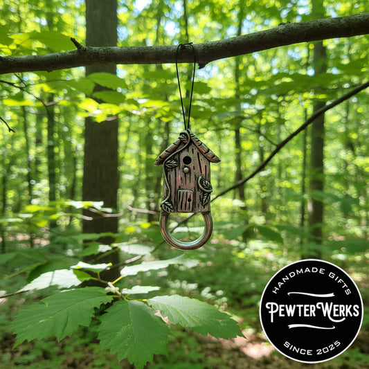 A Pewter 'Birdhouse' Eyeglass Holder hanging from a tree in the woods. PewterWerks logo visible.