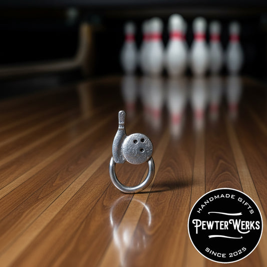 A pewter 'Bowling' eyeglass holder sitting on the hardwood lane in front of the pins. PewterWerks logo visible.