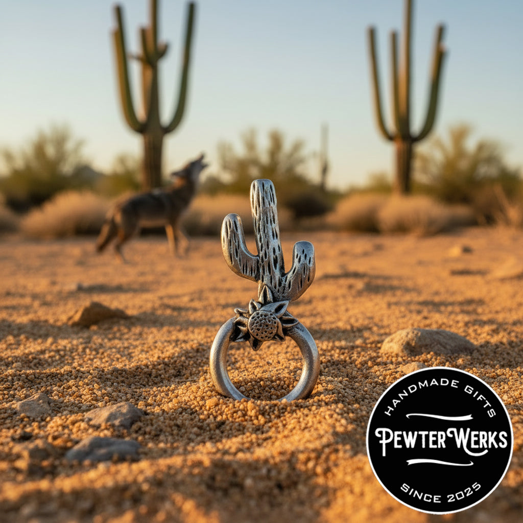 A 'Cactus with Flower' eyeglass holder sitting out in the desert with a coyote howling in the background. PewterWerks logo visible.