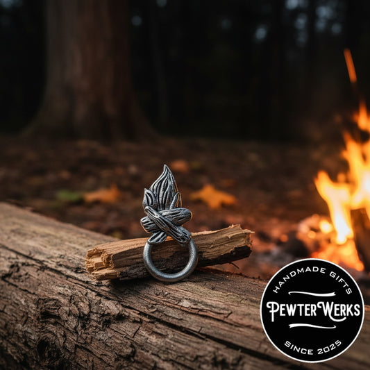 A pewter 'Campfire' eyeglass holder sitting on a log at night next to a campfire. PewterWerks logo visible.