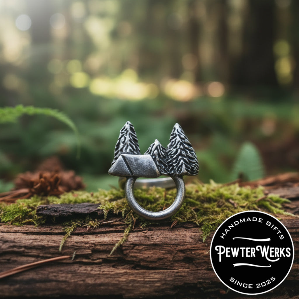 A pewter 'Camping' eyeglass holder sitting on a mossy log in the woods. PewterWerks logo visible.