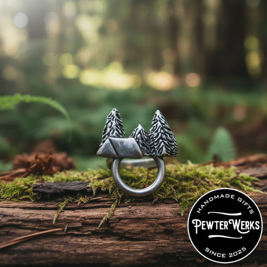 A pewter 'Camping' eyeglass holder sitting on a mossy log in the woods. PewterWerks logo visible.