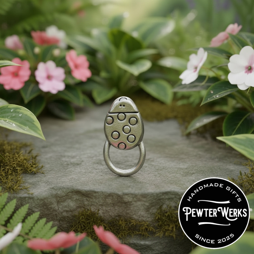 A pewter 'Cartoon Ladybug' eyeglass holder Sitting on a stone ledge amongst plants, flowers and Moss. PewterWerks logo visible.