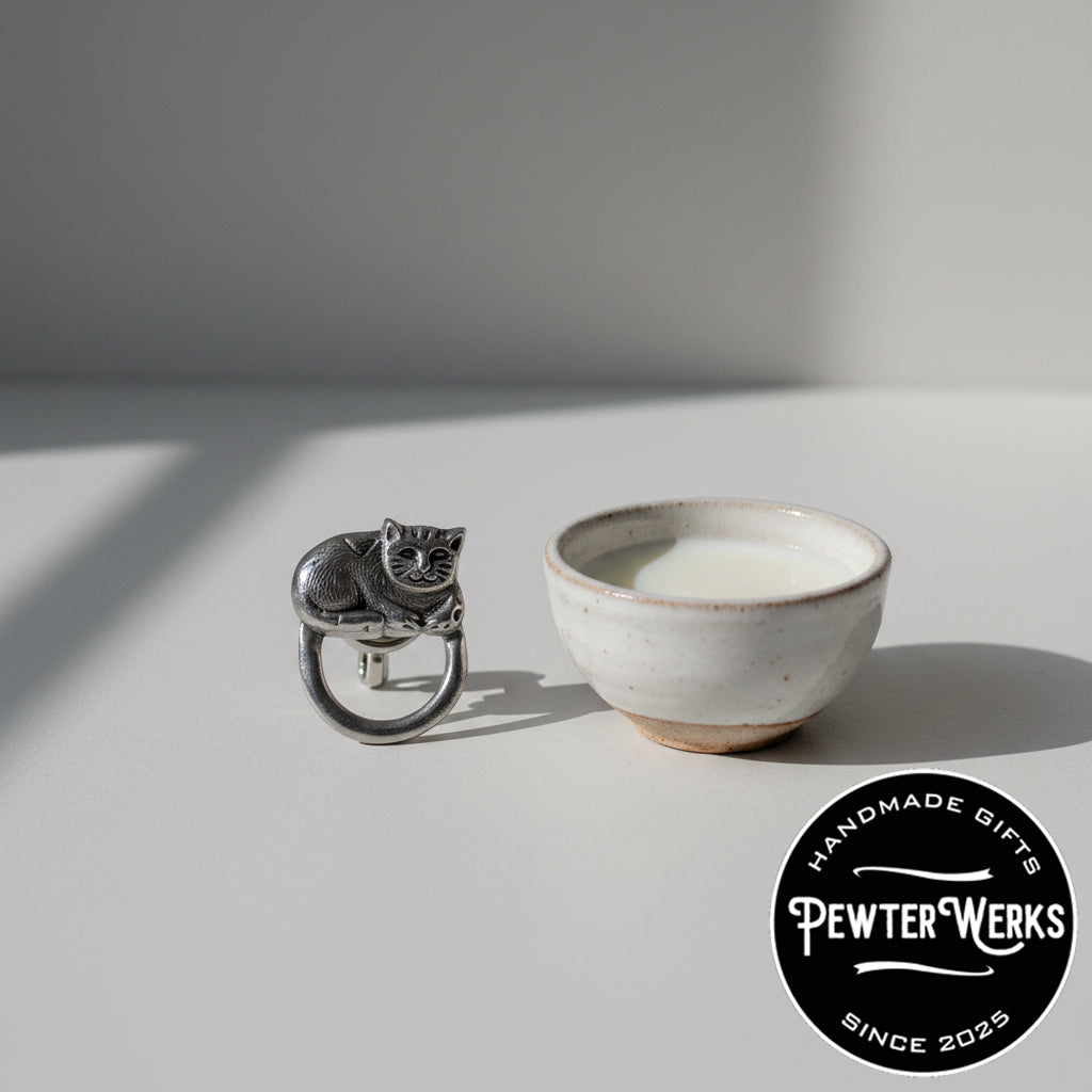 A pewter 'Cat' eyeglass holder next to a bowl of fresh milk. PewterWerks logo is visible.