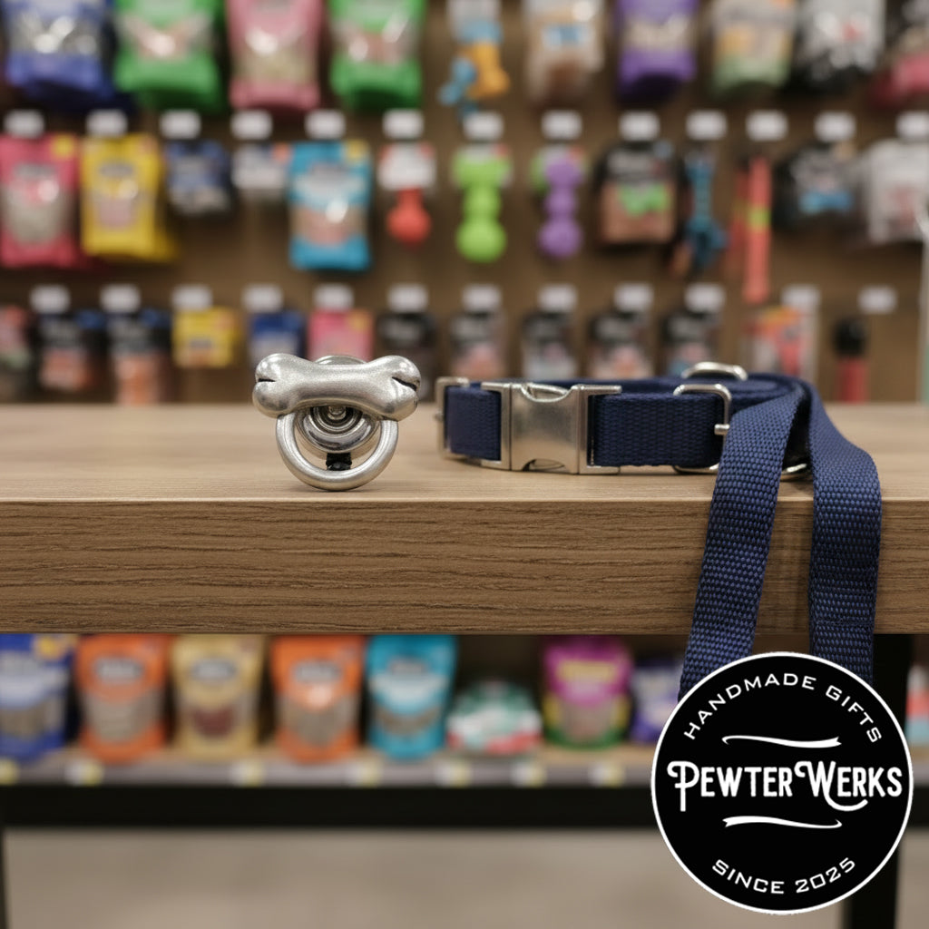 A pewter 'Dog Bone' eyeglass holder sitting on a table next to a dog leash in a local dog shop. PewterWerks logo visible.