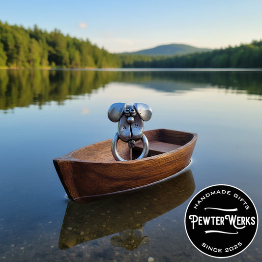 A pewter 'Dog Face No.2' eyeglass holder in a wooden boat out on a beautiful lake. PewterWerks logo visible.