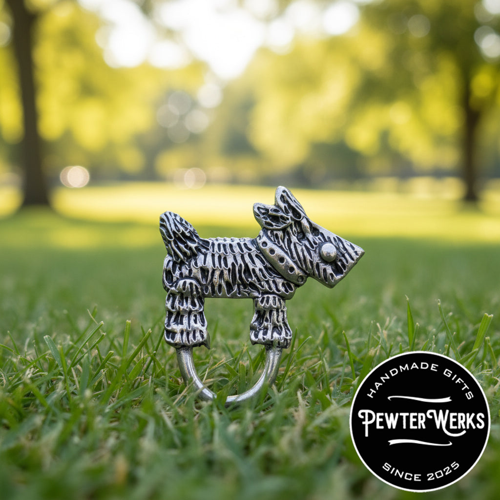 A pewter 'Dog a Scotty' eyeglass holder running around a beautiful green dog park. PewterWerks logo visible.