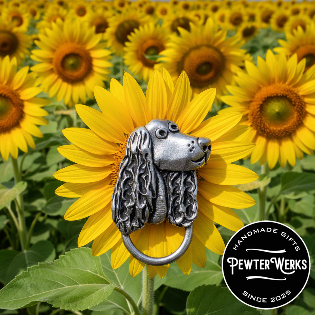 A pewter 'Dog a Spaniel' eyeglass holder hanging out in a field of bright yellow sunflowers. PewterWerks logo visible.