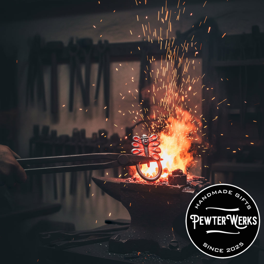 A pewter 'Dragonfly Leprechaun' eyeglass holder being held in the fire with a pair of tongs over an anvil in a blacksmith shop. PewterWerks logo visible.