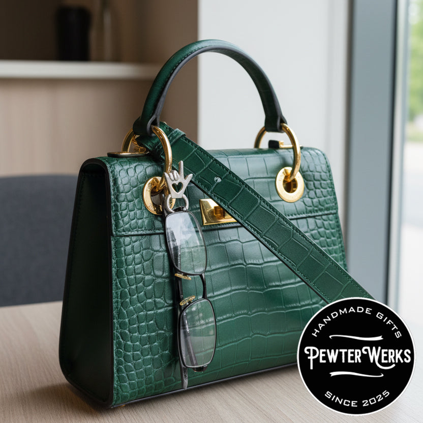A pair of glasses hanging from an 'I Love You' Eyeglass Holder pinned onto a green purse. PewterWerks logo visible.