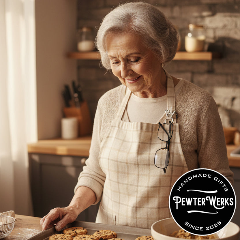 Senior woman baking cookies in her kitchen with her glasses hanging from an 'I Love You' Eyeglass Holder pinned to her apron. PewterWerks logo visible.