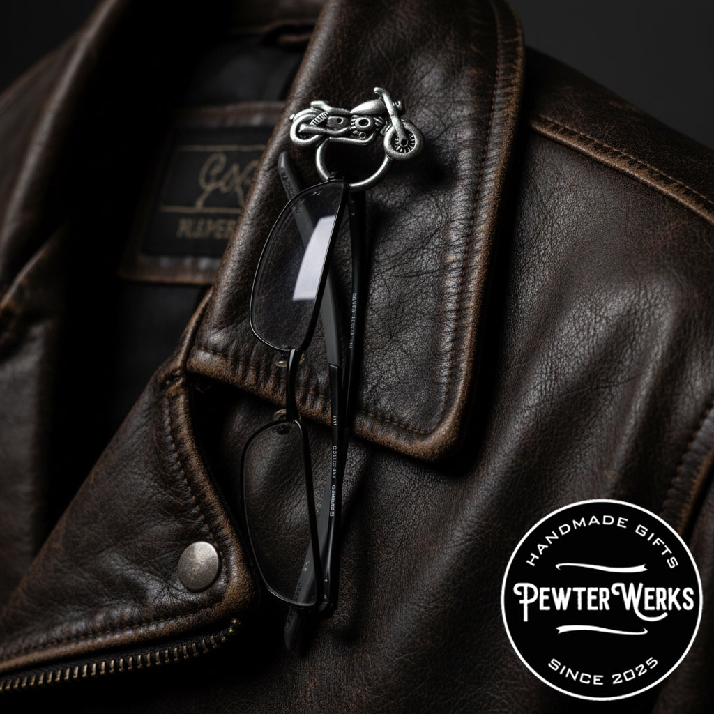 A pair of glasses hanging from 'Motorcycle No.3' Eyeglass Holder pinned to a brown leather jacket. PewterWerks logo visible.